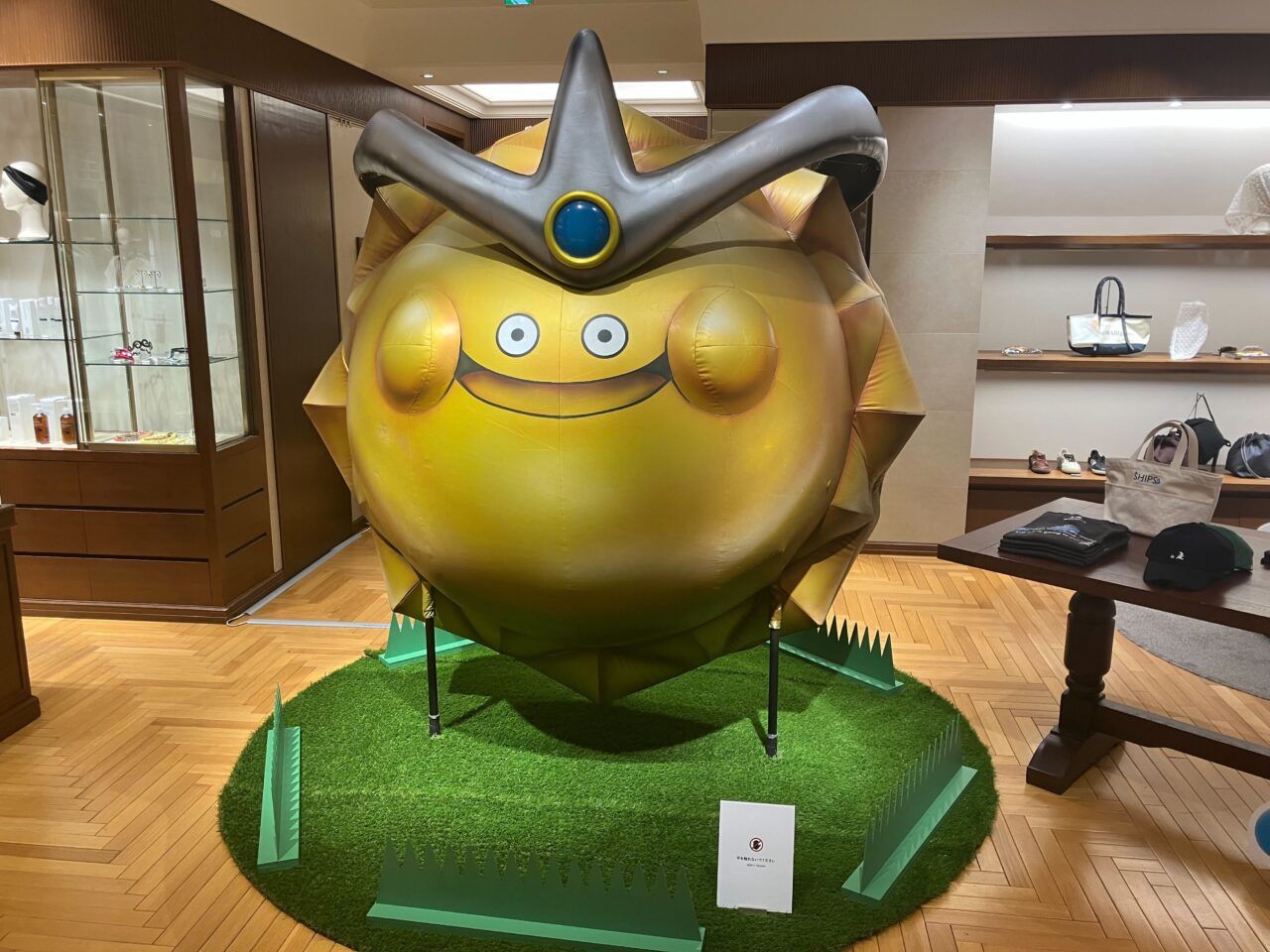 IMG_3903 - innovaTopia Gigantic yellow cartoon mascot indoors, wearing a gray crown-like headpiece with a blue center gem, standing on artificial grass in a store setting.