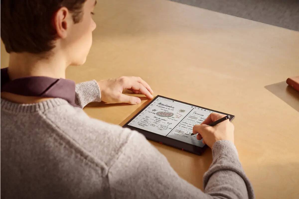 Back view of a person in a gray sweater writing with a stylus on a tablet displaying a menu at a wooden table.
